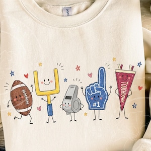 May include: A cream-colored crewneck sweatshirt featuring a graphic print of five anthropomorphic football-themed characters: a brown football, a yellow goalpost, a grey whistle, a blue foam finger with #1, and a maroon pennant labeled TOUCHDOWN.