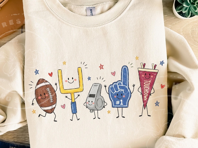 May include: A cream-colored crewneck sweatshirt featuring a graphic print of five anthropomorphic football-themed characters: a brown football, a yellow goalpost, a grey whistle, a blue foam finger with #1, and a maroon pennant labeled TOUCHDOWN.