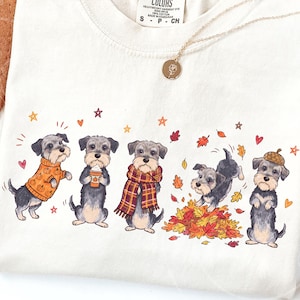 Puede incluir: Una camiseta de color crema con un estampado gráfico de cinco perros schnauzer grises y negros con atuendos otoñales, incluyendo un suéter de calabazas, una bufanda de cuadros y un sombrero de bellota, rodeados de hojas de otoño, estrellas y corazones.