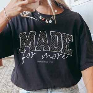 Puede incluir: Camiseta negra con las palabras "MADE for more" en fuente blanca. Las letras "MADE" tienen un dise&ntilde;o de estampado de leopardo gris. El texto "Ephesians 2:10" est&aacute; debajo de las palabras "for more".