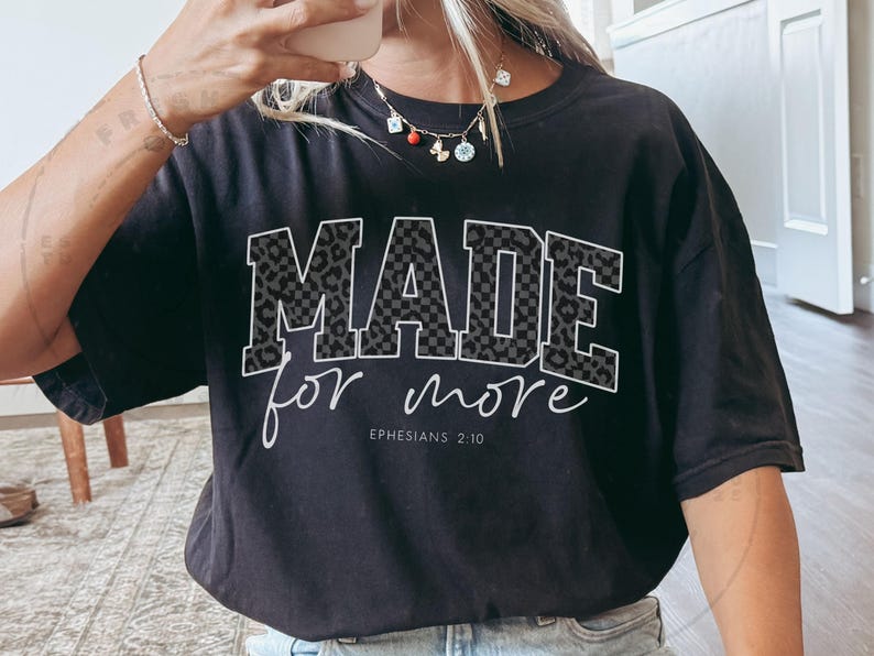 Puede incluir: Camiseta negra con las palabras "MADE for more" en fuente blanca. Las letras "MADE" tienen un dise&ntilde;o de estampado de leopardo gris. El texto "Ephesians 2:10" est&aacute; debajo de las palabras "for more".