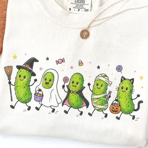 Puede incluir: Una camiseta de color crema con un gráfico de cinco pepinillos verdes disfrazados de Halloween, incluyendo una bruja, un fantasma, un vampiro, una momia y un gato, acompañados de ilustraciones de dulces coloridos y estrellas.