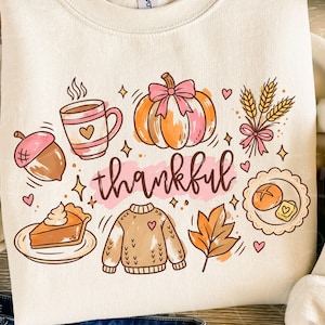 May include: A cream-coloured crewneck sweatshirt featuring a colourful graphic print with the word thankful in cursive script. The design includes a pumpkin with a pink bow, a coffee mug, an acorn, a slice of pie, a sweater, wheat, and a leaf.
