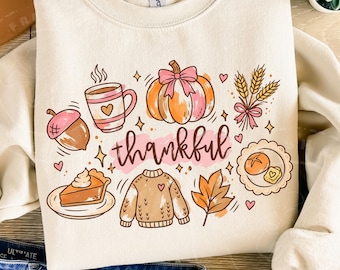 May include: A cream-coloured crewneck sweatshirt featuring a colourful graphic print with the word thankful in cursive script. The design includes a pumpkin with a pink bow, a coffee mug, an acorn, a slice of pie, a sweater, wheat, and a leaf.