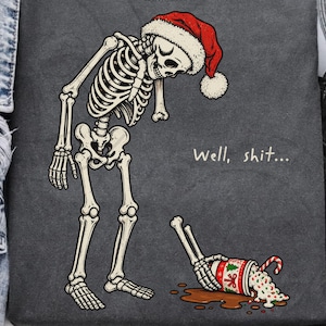 May include: A dark gray t-shirt featuring a graphic of a skeleton wearing a red Santa hat, looking down at a spilled cup of hot cocoa with a candy cane. The text "Well, shit..." is printed in white next to the skeleton.