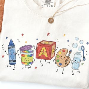 May include: A cream-colored t-shirt featuring a graphic print of five anthropomorphic school supplies: a blue crayon, a rainbow-filled paint container, a red alphabet block with the letter A, a wooden paint palette with a brush, and a bubble wand.