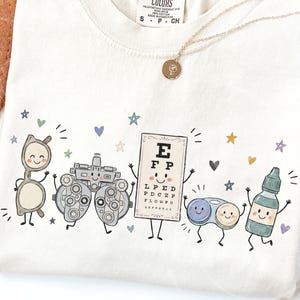 May include: A cream-colored t-shirt featuring a graphic print of five anthropomorphic eye care items: a pair of glasses, a phoropter, an eye chart with the letters E, F, P, L, P, E, D, P, D, C, Z, P, F, L, O, W, P, C, contact lens case, and eye drops.