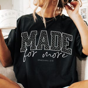 Puede incluir: Camiseta negra con las palabras "MADE for more" en un dise&ntilde;o blanco, de leopardo y a cuadros. El texto "Efesios 2:10" est&aacute; debajo. La camiseta es de estilo casual.