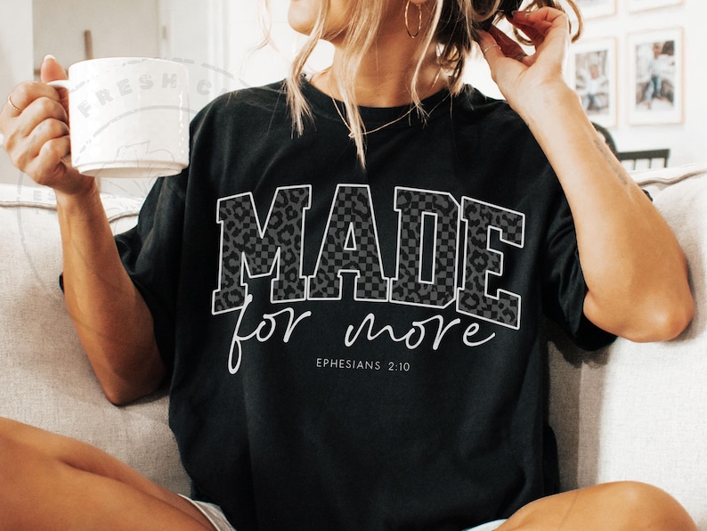 Puede incluir: Camiseta negra con las palabras "MADE for more" en un dise&ntilde;o blanco, de leopardo y a cuadros. El texto "Efesios 2:10" est&aacute; debajo. La camiseta es de estilo casual.