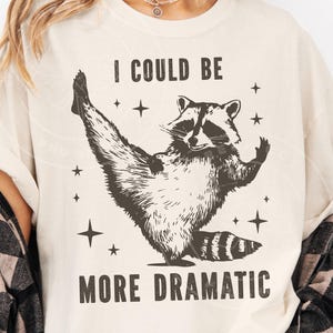Puede incluir: Camiseta color crema con una ilustración de mapache en tonos marrón y negro, con el texto "I COULD BE MORE DRAMATIC". El mapache está en una pose juguetona, rodeado de estrellas.