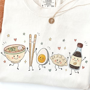 Könnte beinhalten: Ein cremefarbenes T-Shirt mit einer verspielten Grafik aus anthropomorphen Lebensmitteln, darunter eine Schüssel Ramen, Stäbchen, ein gekochtes Ei, ein Dumpling und eine Sojasaucenflasche, alle mit lächelnden Gesichtern und kleinen Sternen und Herzen.