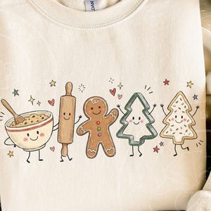 May include: A cream-colored crewneck sweatshirt featuring a colorful graphic print of anthropomorphic baking items including a mixing bowl with a wooden spoon, a rolling pin, a gingerbread person, and two decorated Christmas tree cookies.