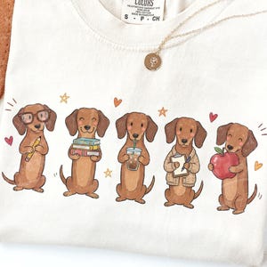 May include: A cream-colored t-shirt featuring a graphic of five brown dachshunds. The dogs are depicted holding a yellow pencil, a stack of books, an iced coffee, a clipboard, and a red apple, surrounded by small orange hearts and stars.