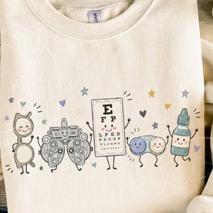 May include: A cream-colored crewneck sweatshirt featuring a graphic print of five anthropomorphic eye care items: a pair of glasses, a phoropter, an eye chart with the letters E, F, P, L, P, E, D, P, D, C, Z, P, F, L, O, W, F, O, a contact lens case, and an eye drop bottle.