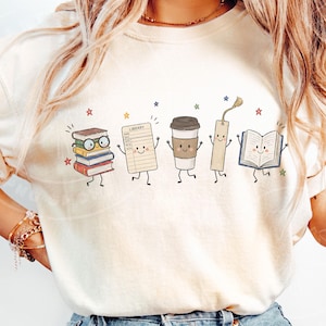 Pu&ograve; includere: Una t-shirt color crema con una stampa grafica di cinque personaggi antropomorfi: una pila di libri con occhiali, una scheda di biblioteca con la parola LIBRARY, un bicchiere di caff&egrave;, un segnalibro e un libro aperto, circondati da stelle.
