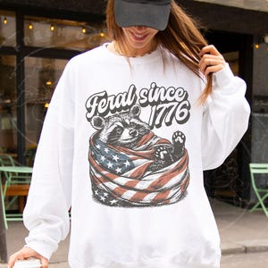 May include: White sweatshirt featuring a raccoon wrapped in an American flag. The text "Feral since 1776" is arched above the raccoon. The sweatshirt has long sleeves and a crew neck.