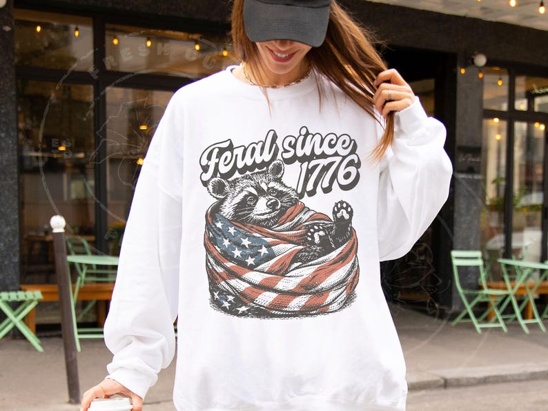 May include: White sweatshirt featuring a raccoon wrapped in an American flag. The text "Feral since 1776" is arched above the raccoon. The sweatshirt has long sleeves and a crew neck.
