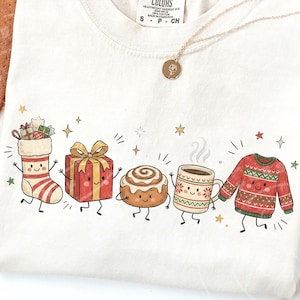 May include: A cream-coloured t-shirt featuring a graphic print of five anthropomorphic holiday items: a striped stocking, a red gift box with a gold bow, a cinnamon roll, a steaming mug, and a red and green patterned jumper, all with faces.