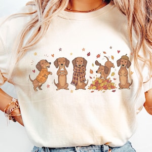 May include: A cream-colored t-shirt featuring a graphic print of five brown dachshunds in autumn-themed attire and activities, including wearing a pumpkin sweater, holding a coffee cup, wearing a plaid scarf, playing in leaves, and wearing an acorn hat.