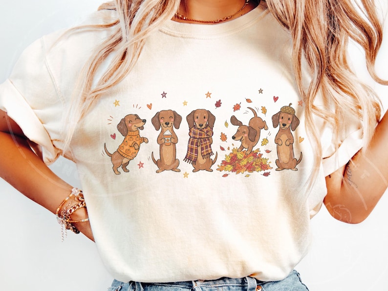May include: A cream-colored t-shirt featuring a graphic print of five brown dachshunds in autumn-themed attire and activities, including wearing a pumpkin sweater, holding a coffee cup, wearing a plaid scarf, playing in leaves, and wearing an acorn hat.
