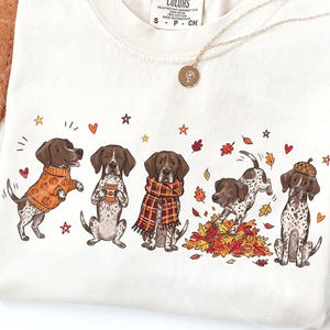 May include: An off-white t-shirt featuring a graphic print of five brown and white spotted dogs. The dogs are depicted wearing autumn-themed accessories like sweaters, scarves, and hats, surrounded by falling leaves, stars, and small hearts.