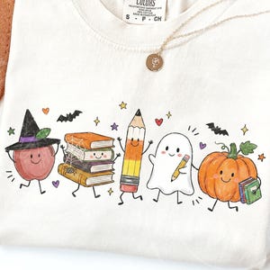 May include: A cream-colored t-shirt featuring a colorful graphic of a smiling apple wearing a witch hat, a stack of books with a spider web, a pencil, a ghost holding a pencil, and a pumpkin holding a book, accented with bats and stars.