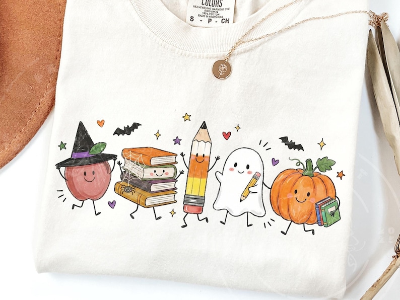 May include: A cream-colored t-shirt featuring a colorful graphic of a smiling apple wearing a witch hat, a stack of books with a spider web, a pencil, a ghost holding a pencil, and a pumpkin holding a book, accented with bats and stars.