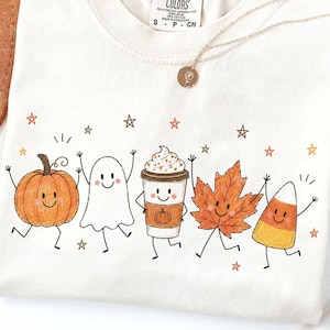May include: Cream-colored sweatshirt featuring a whimsical fall-themed graphic. The design includes a smiling pumpkin, ghost, coffee cup, maple leaf, and candy corn, all dancing with joy. Gold necklace with a pendant.