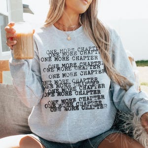 K&ouml;nnte beinhalten: Hellgraues Sweatshirt mit dem Text "ONE MORE CHAPTER" in Schwarz. Der Text wiederholt sich mehrfach auf der Vorderseite des Sweatshirts. Die Person h&auml;lt ein Glas eines braunen Getr&auml;nks.