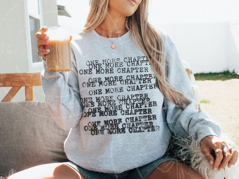 K&ouml;nnte beinhalten: Hellgraues Sweatshirt mit dem Text "ONE MORE CHAPTER" in Schwarz. Der Text wiederholt sich mehrfach auf der Vorderseite des Sweatshirts. Die Person h&auml;lt ein Glas eines braunen Getr&auml;nks.