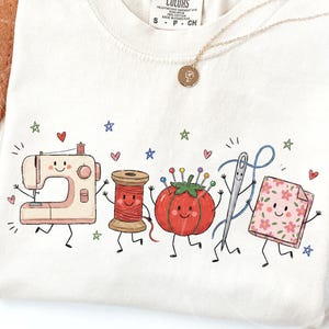 Könnte beinhalten: Ein cremefarbenes T-Shirt mit einer bunten Grafik von fünf anthropomorphen Nähutensilien: eine beige Nähmaschine, eine rote Garnrolle, ein rotes Tomaten-Nadelkissen, eine silberne Nähnadel mit blauem Faden und ein rosa Stoffquadrat mit Blumenmuster.