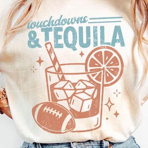 Puede incluir: Una camiseta de color crema con un estampado gráfico que incluye el texto "touchdowns & TEQUILA" en azul. El diseño muestra un vaso de cóctel con cubitos de hielo, una pajita a rayas, una rodaja de cítrico y un balón de fútbol americano naranja.