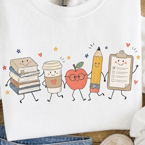 Peut inclure: Un sweat-shirt blanc &agrave; col rond orn&eacute; d'un motif color&eacute; repr&eacute;sentant cinq fournitures scolaires anthropomorphes se tenant par la main. Les personnages incluent une pile de livres, un gobelet &agrave; caf&eacute;, une pomme &agrave; lunettes, un crayon et un presse-papier.