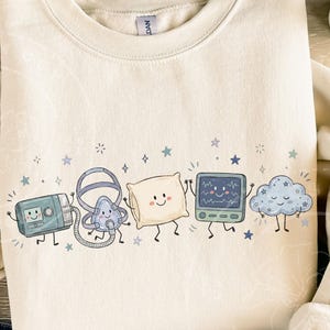 May include: A cream-colored crewneck sweatshirt featuring a graphic print of five anthropomorphic characters: a CPAP machine, a nasal mask, a pillow, a heart rate monitor, and a cloud, all decorated with small blue stars and sparkles.