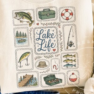 May include: Cream-colored sweatshirt featuring a collection of fishing-themed illustrations. The design includes a fish, tackle box, life preserver, fishing rod, boat, lure, hat, worm, dock, and the words "Lake Life".
