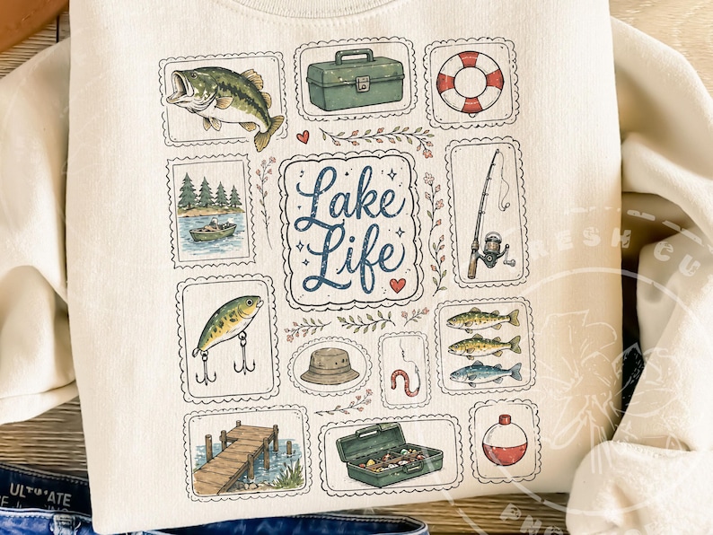 May include: Cream-colored sweatshirt featuring a collection of fishing-themed illustrations. The design includes a fish, tackle box, life preserver, fishing rod, boat, lure, hat, worm, dock, and the words "Lake Life".