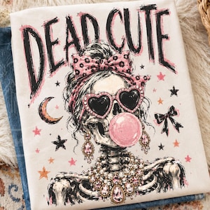May include: A cream-colored t-shirt featuring a graphic of a skeleton wearing heart-shaped sunglasses, a leopard print bow, and a jeweled necklace, blowing a pink bubble. The text DEAD CUTE is printed in black above the skeleton with pink accents.