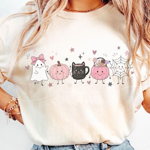 May include: A cream-colored t-shirt featuring a graphic print of five cute characters: a white ghost with a pink bow, a pink pumpkin, a black mug, a pink pumpkin bucket filled with candy, and a spiderweb, all accented by small stars and hearts.