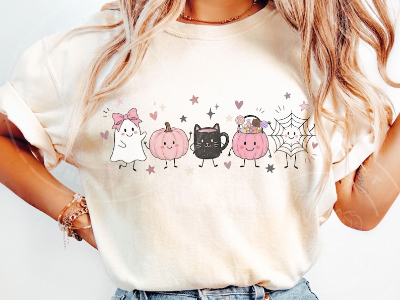 May include: A cream-colored t-shirt featuring a graphic print of five cute characters: a white ghost with a pink bow, a pink pumpkin, a black mug, a pink pumpkin bucket filled with candy, and a spiderweb, all accented by small stars and hearts.