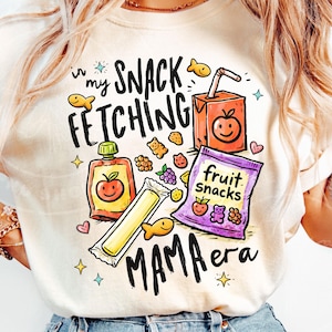 May include: A cream-coloured t-shirt with a vibrant graphic. The design includes the text "in my snack fetching mama era" alongside illustrations of snacks such as juice boxes, fruit snacks, and a fruit pouch. Fish-shaped crackers and other food items are also depicted.