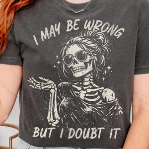 Könnte beinhalten: Dunkelgraues T-Shirt mit einem Skelett-Motiv, das eine Sonnenbrille und einen Dutt trägt. Der Text lautet "I MAY BE WRONG BUT I DOUBT IT" in Weiß. Das Shirt hat einen Vintage-Look.