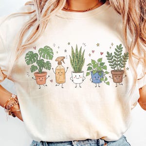 K&ouml;nnte beinhalten: Ein cremefarbenes T-Shirt mit einem Grafikdruck von f&uuml;nf anthropomorphen Charakteren: eine Monstera im Terrakottatopf, eine Spr&uuml;hflasche, eine Bogenhanfpflanze im wei&szlig;en Topf, eine Efeutute im blauen Topf und eine Gl&uuml;cksfeder im braunen Topf.