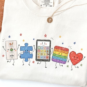 May include: A cream-colored t-shirt featuring a graphic of five animated characters: a white communication board with snack, read, and play icons, a blue puzzle piece, a tablet with a smiley face, a rainbow pop-it toy, and a red heart, all with limbs.