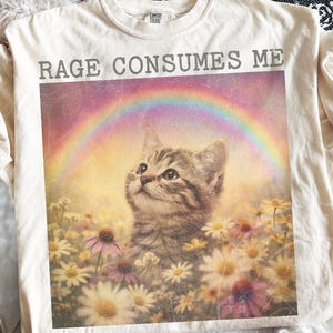 May include: Cream-colored t-shirt featuring a graphic of a kitten looking up at a rainbow, surrounded by flowers. The text "RAGE CONSUMES ME" is printed above the graphic in gray letters. The shirt has a crew neck and short sleeves.