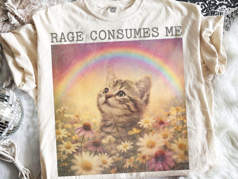 May include: Cream-colored t-shirt featuring a graphic of a kitten looking up at a rainbow, surrounded by flowers. The text "RAGE CONSUMES ME" is printed above the graphic in gray letters. The shirt has a crew neck and short sleeves.