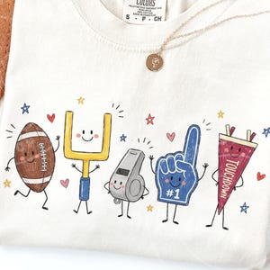 May include: An off-white t-shirt featuring a graphic print of five anthropomorphic football-themed characters: a brown football, a yellow goalpost, a grey whistle, a blue foam finger with the text #1, and a red pennant with the text TOUCHDOWN.