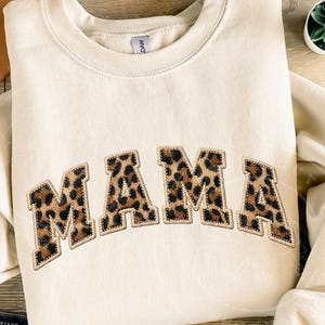 Op de afbeelding: Crèmekleurige sweatshirt met het woord "MAMA" in luipaardprint letters. De letters zijn gebogen en hebben een gestikte rand. De sweatshirt is opgevouwen, met een kleine vetplant zichtbaar op de achtergrond.