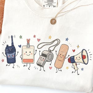 May include: A cream-colored t-shirt featuring a graphic print of five anthropomorphic characters: a dark blue walkie-talkie, a white ID badge, a silver whistle, a tan adhesive bandage, and a red, white, and blue megaphone, all with smiling faces.