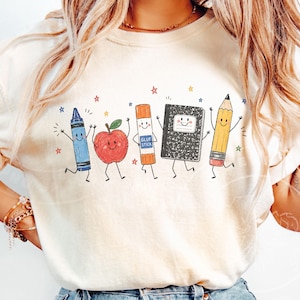 May include: A cream-coloured t-shirt with a cheerful design of animated school supplies. A blue crayon, red apple, glue stick, notebook, and pencil are shown with smiling faces and dancing, with colourful stars around them.