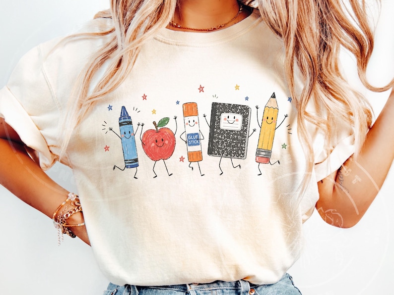 May include: A cream-coloured t-shirt with a cheerful design of animated school supplies. A blue crayon, red apple, glue stick, notebook, and pencil are shown with smiling faces and dancing, with colourful stars around them.